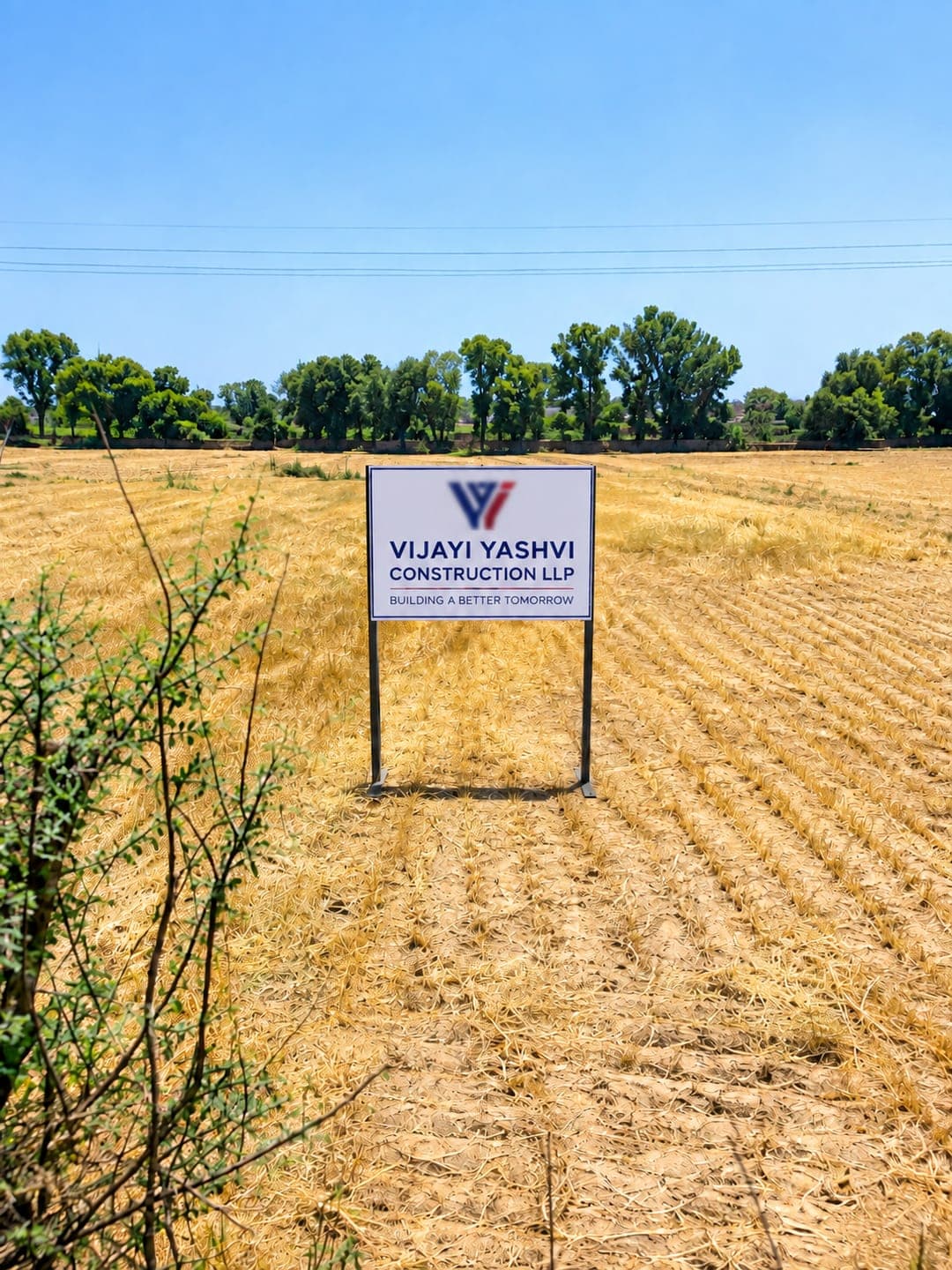 A site board reading "Vijayi Yashvi Construction LLP, Building a Better Tomorrow" standing in a harvested field, with a tree line along the far boundary and power lines overhead.